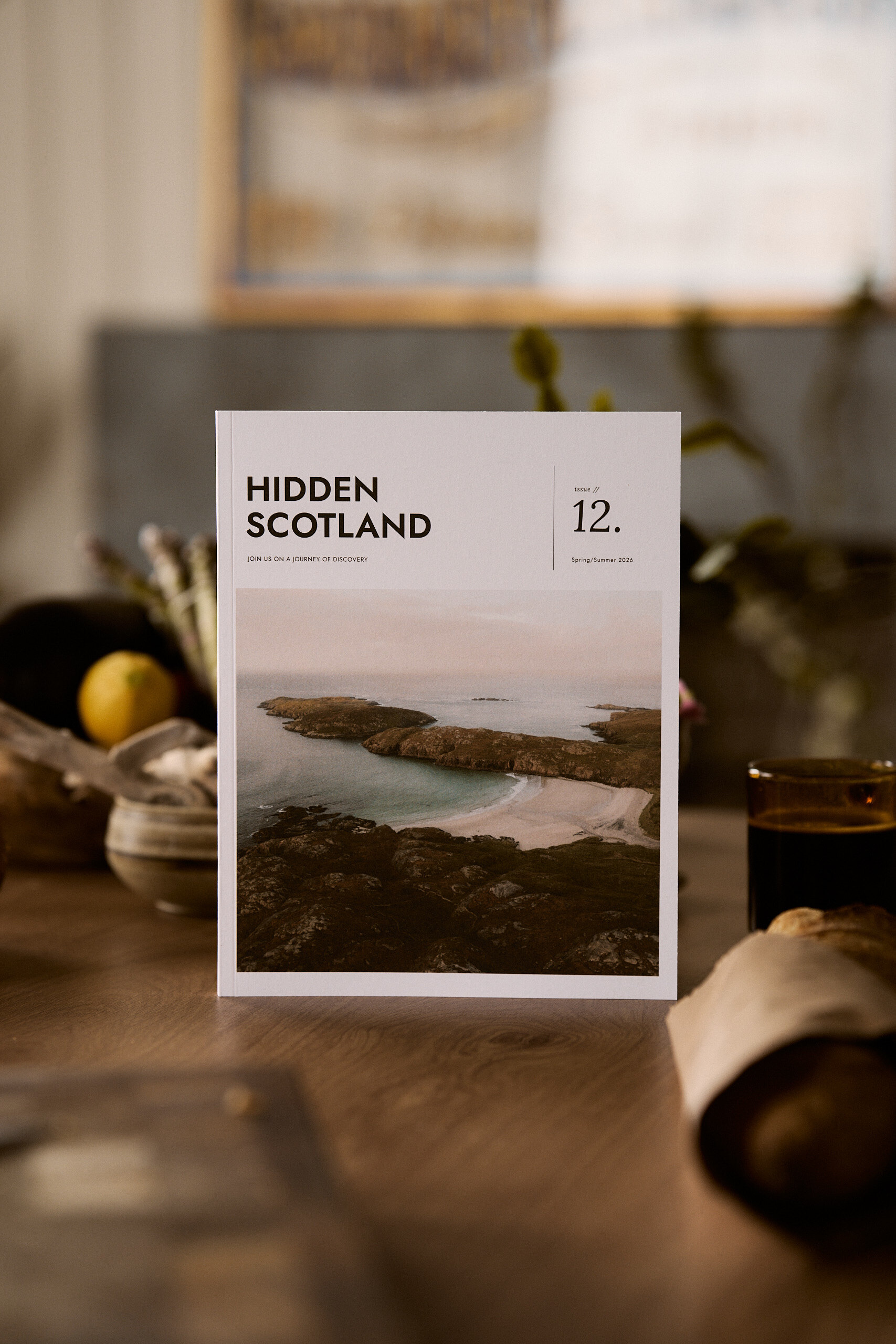A copy of Hidden Scotland issue 12 showing a coastal cliff and sandy bay on its cover stands upright on a wooden table beside a lemon, driftwood and a glass of dark liquid, a framed picture blurred behind.