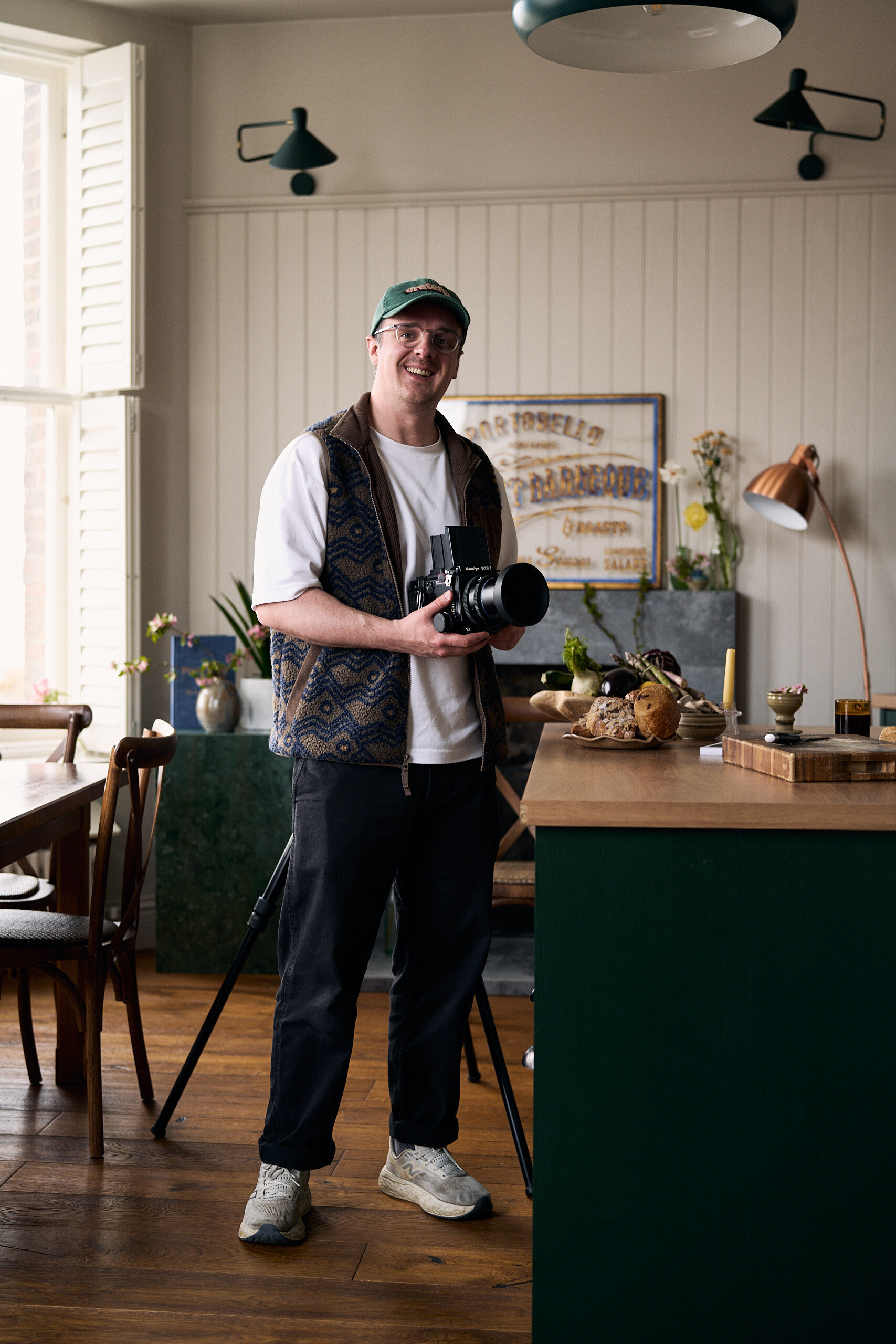 A man in a patterned gilet and green cap smiles while holding a camera in a green-walled dining room, a framed vintage advertisement and a lit lamp behind him.