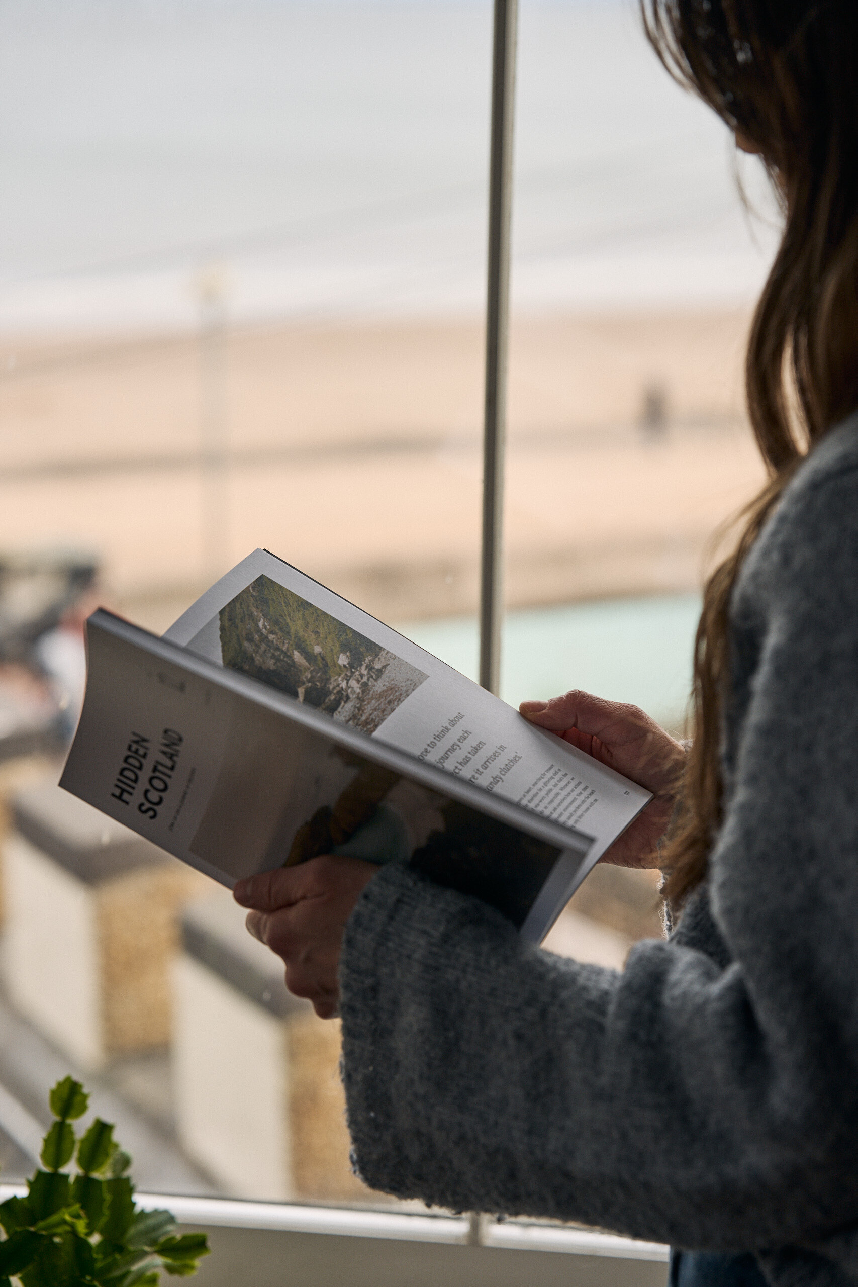 A woman reads a magazine beside a window overlooking a sandy beach, her reflection faint in the glass.