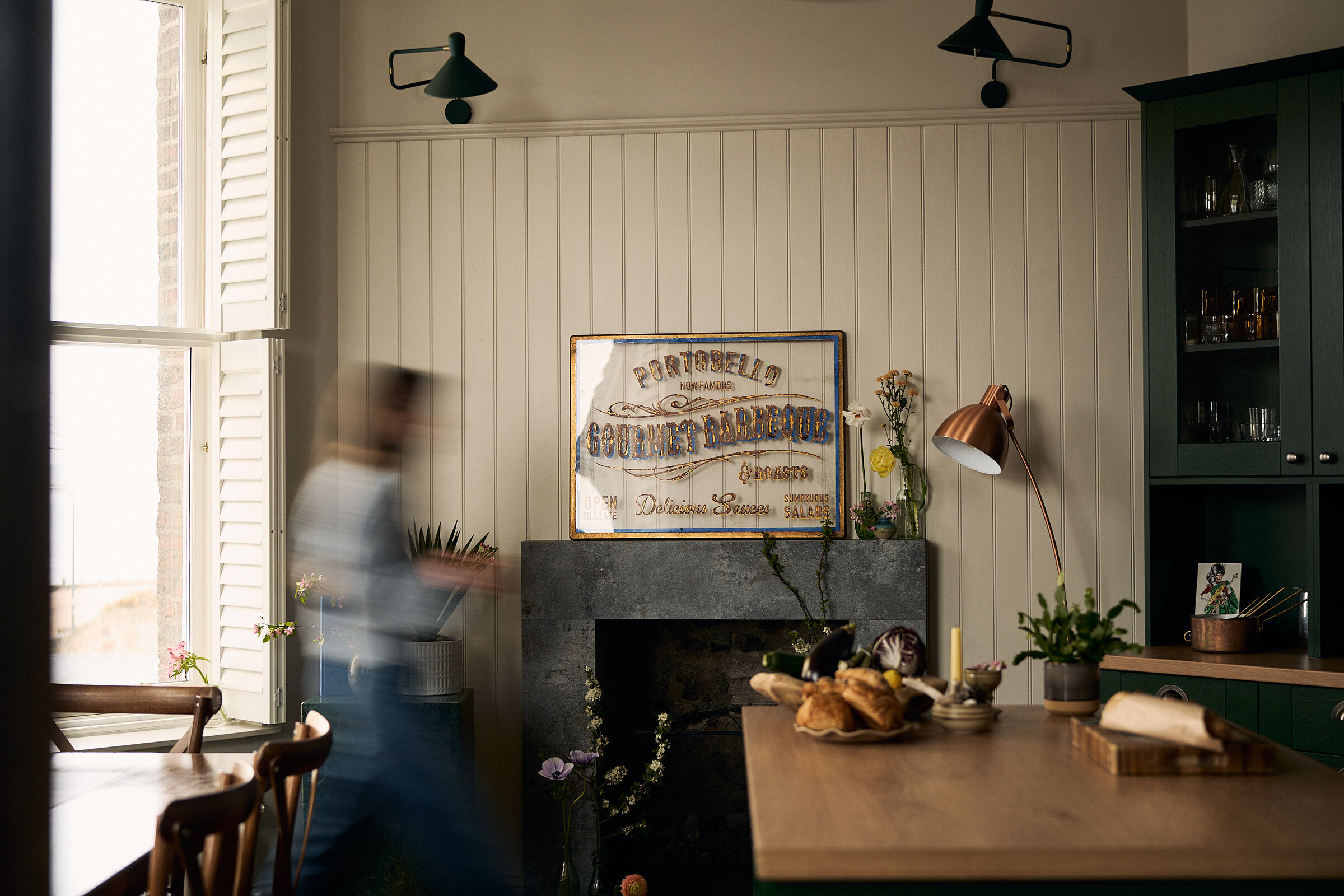 A woman in motion blur walks past a fireplace beneath a framed vintage advertisement sign, a lamp and a glass-fronted dresser in a green-panelled dining room.