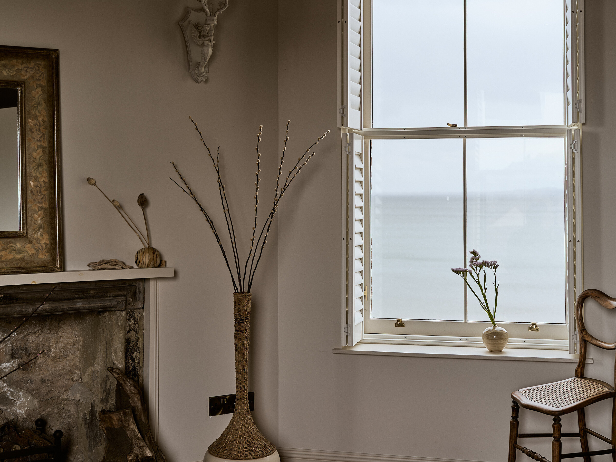 A tall vase of pussy willow branches stands beside a gilt-framed mirror and a marble fireplace, a sash window overlooking the sea and a small vase of flowers on the sill.