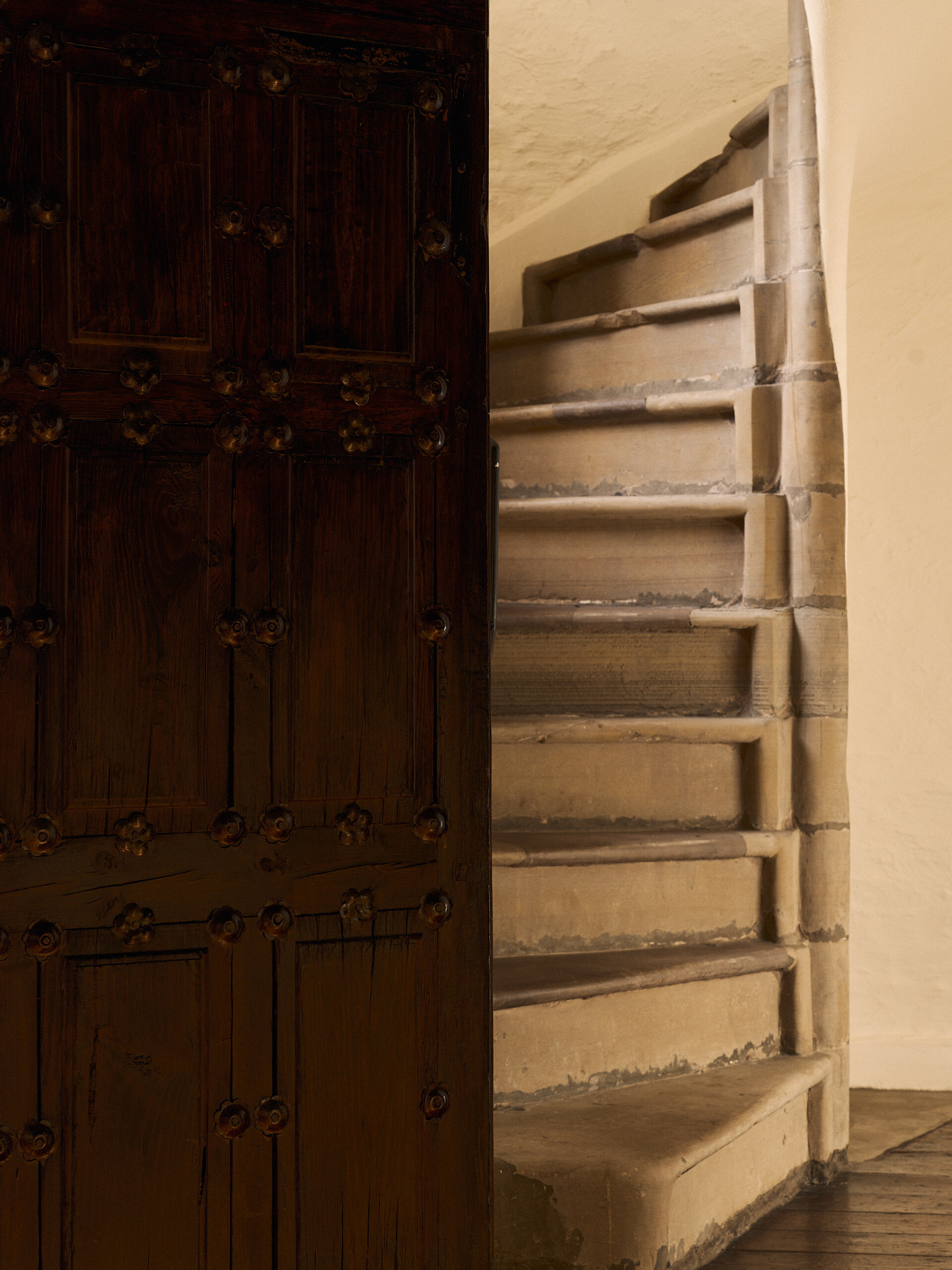 A stone spiral staircase curves upward beside a heavy studded wooden door, warm plastered walls surrounding it.