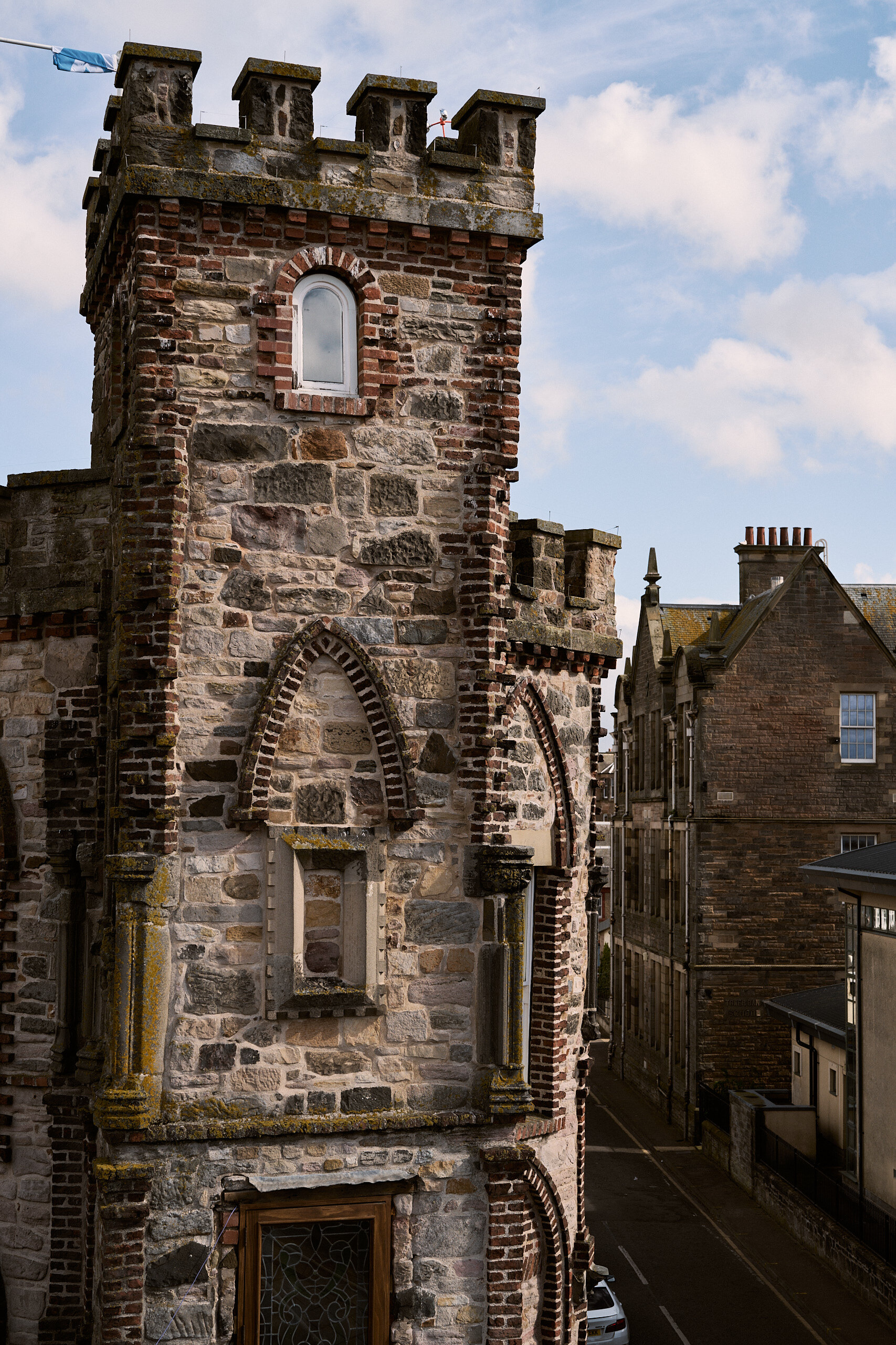 A stone castellated tower with an arched window and red brick trim rises above a street of sandstone tenements and chimney pots.