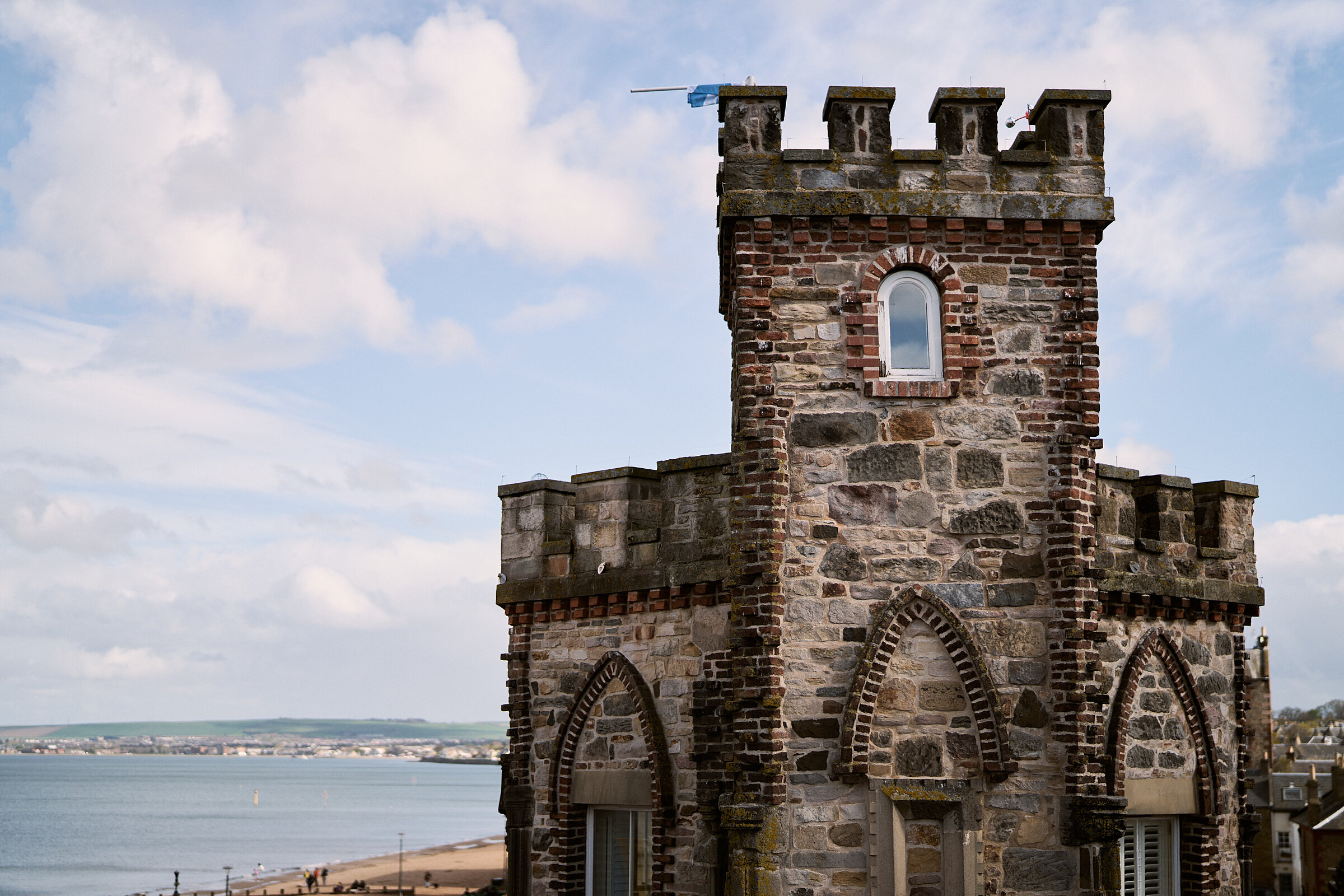 Crenellated stone tower rising above a coastal town, with a bay and shoreline stretching into the distance