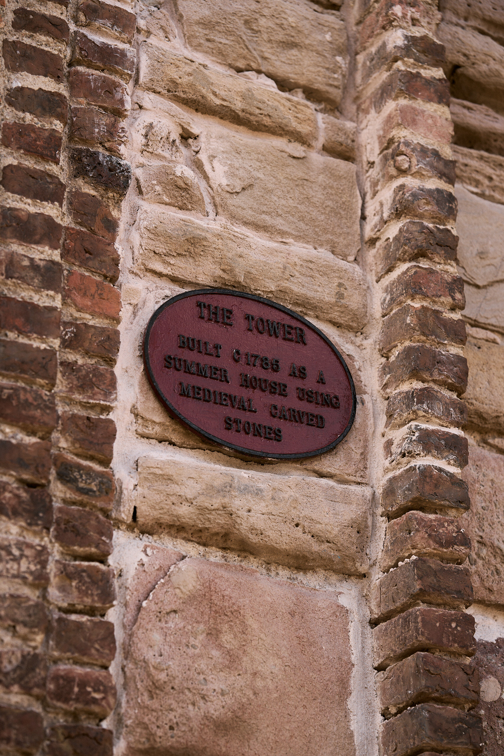 An oval plaque set in a stone wall reads The Tower, built around 1866 as a summer house using medieval carved stones