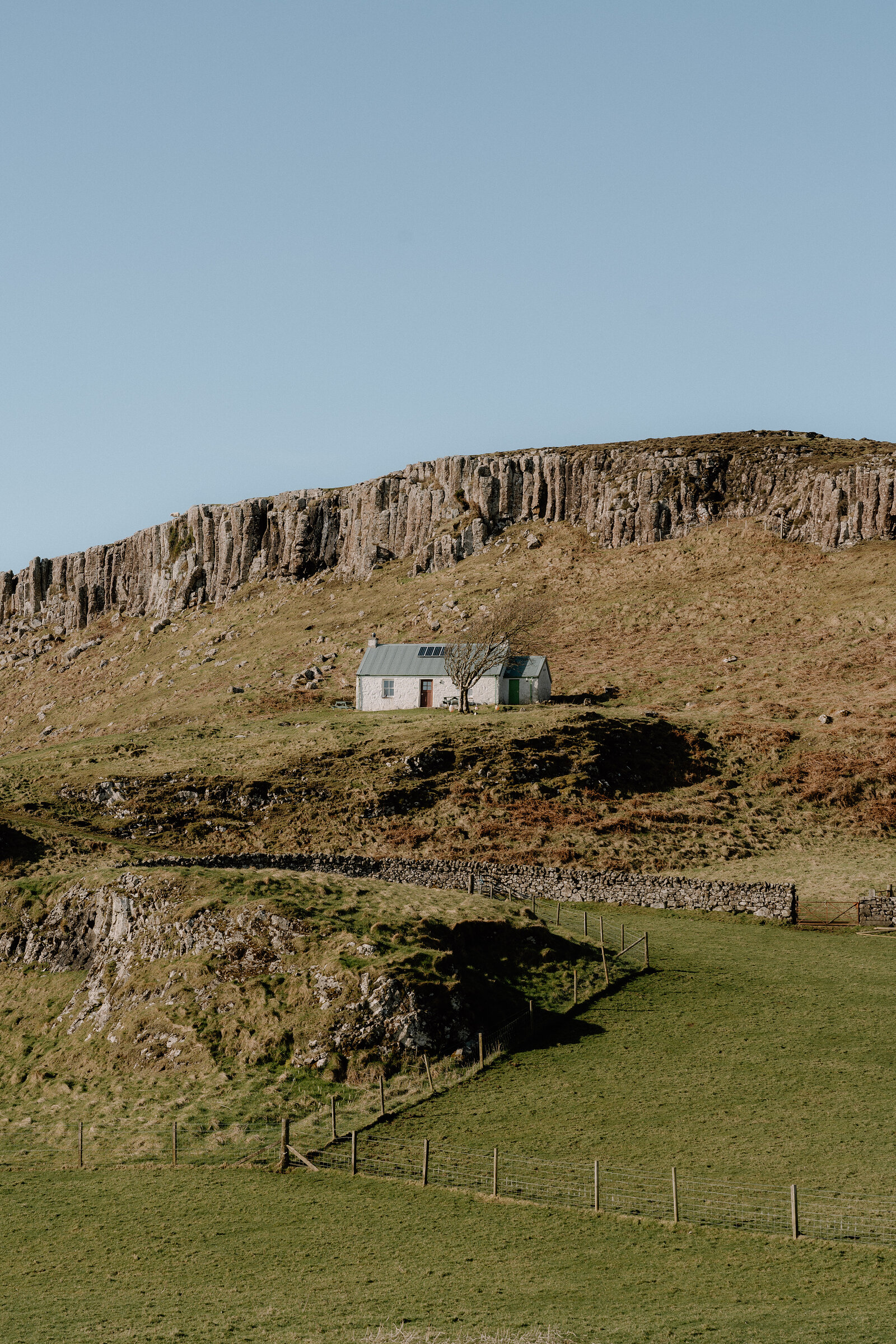A white cottage with a corrugated roof sits beneath columnar basalt cliffs on a grassy hillside.