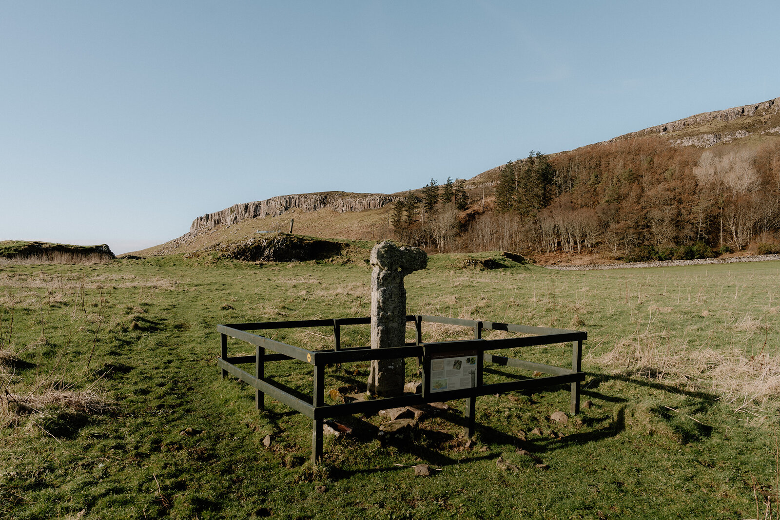 A protective fence surrounds the Isle of Canna's ancient stone cross in a grassy field.