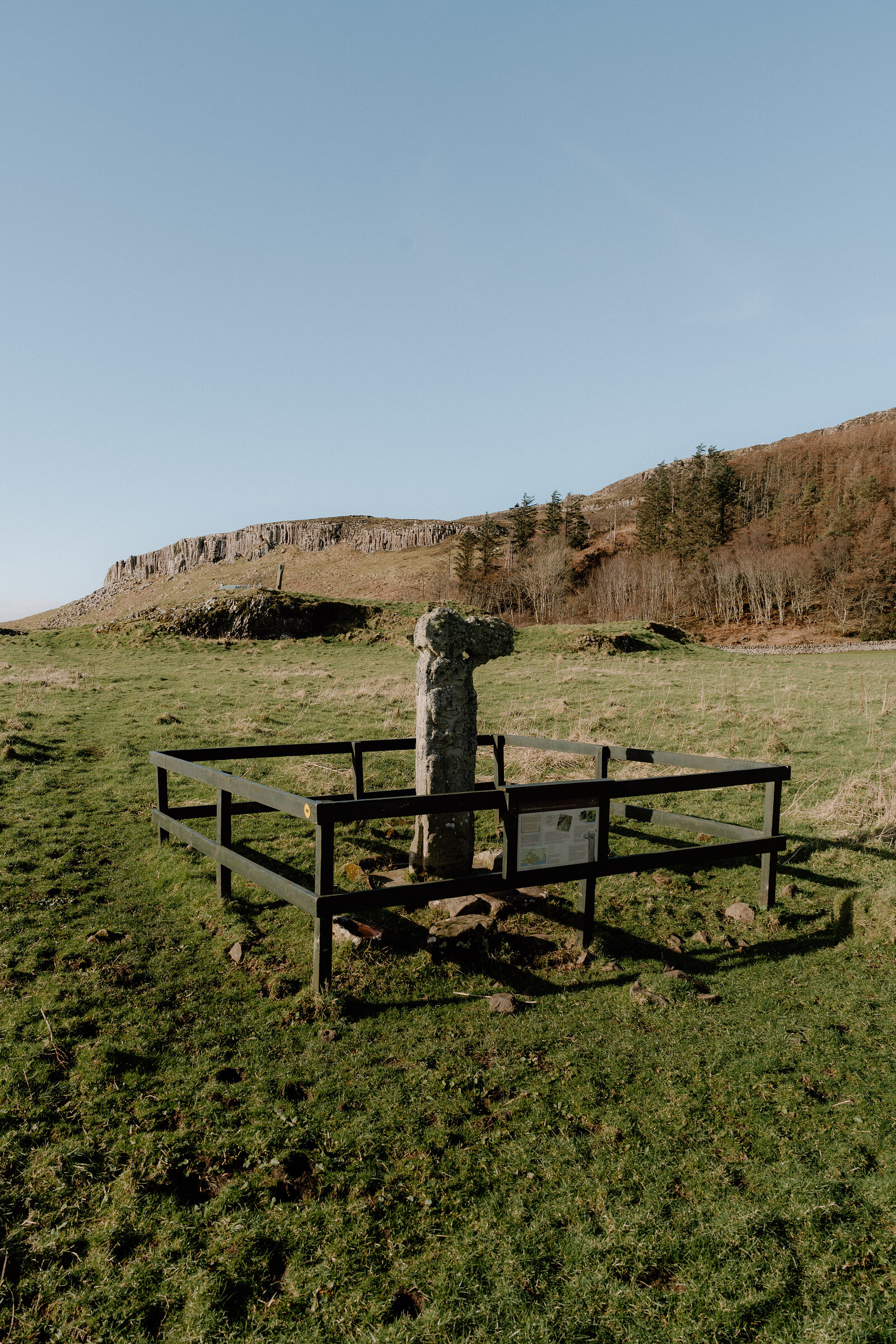 An interpretive sign stands beside a fenced stone cross in a field beneath wooded cliffs on the Isle of Canna.