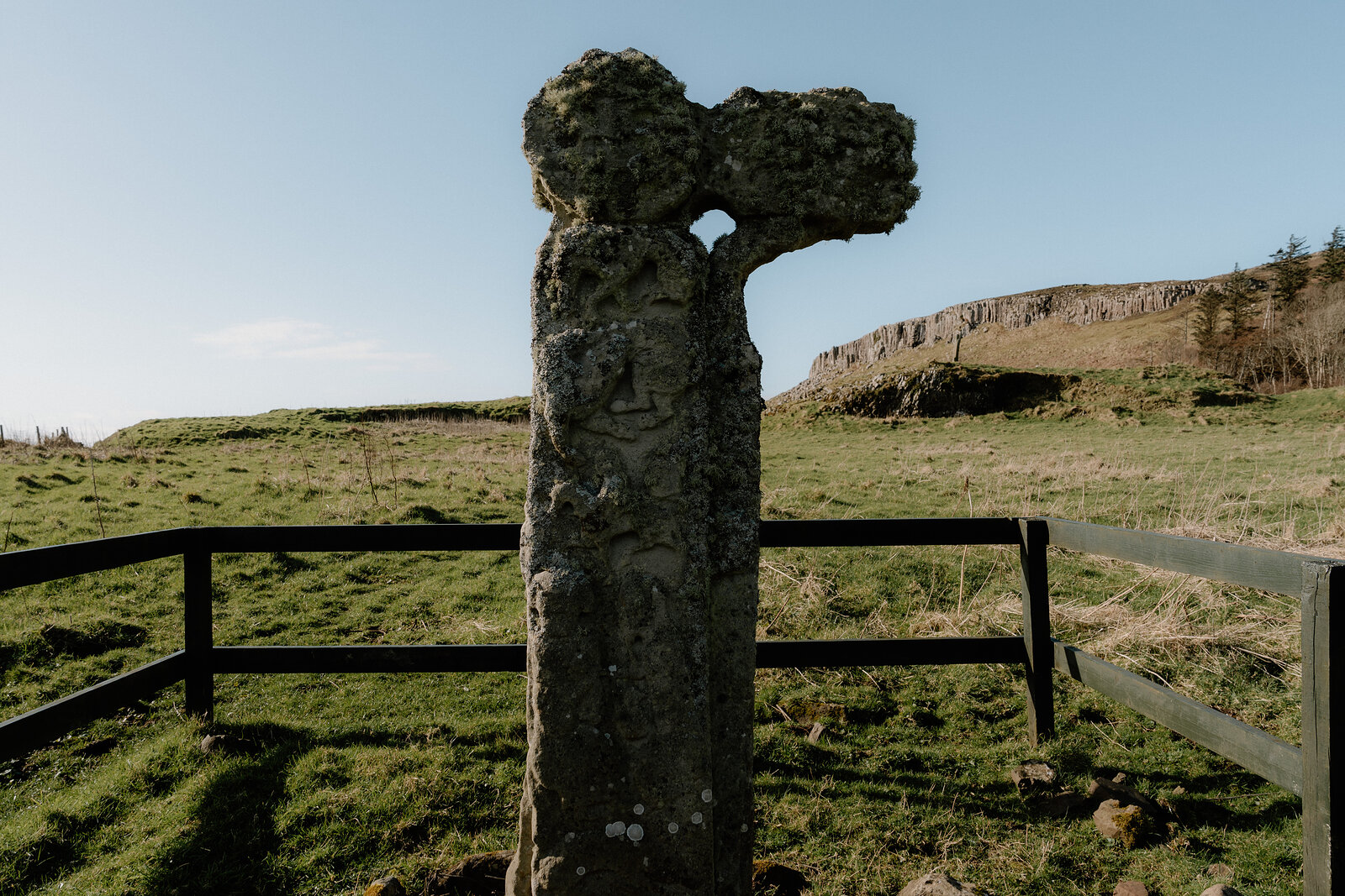 The Isle of Canna's ancient stone cross stands within a post-and-rail fence before a rocky ridge.