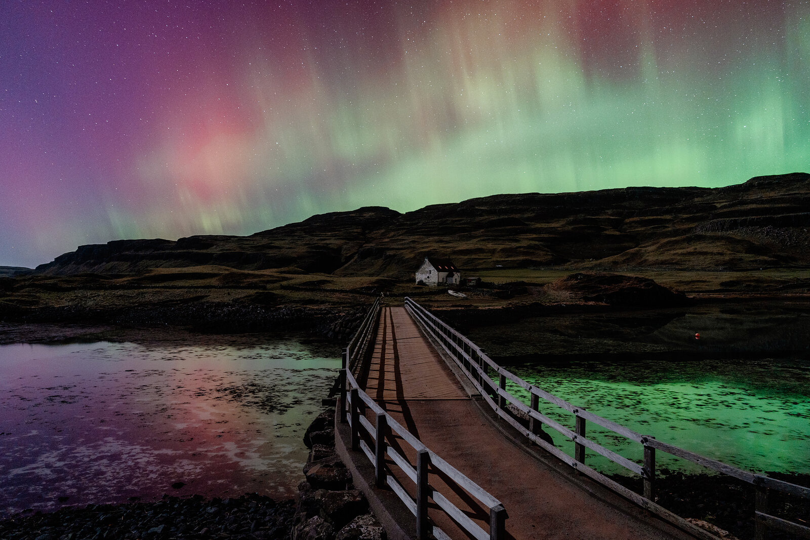 Pink and green Aurora Borealis fills the sky above a wooden footbridge leading to a cottage on the Isle of Canna.