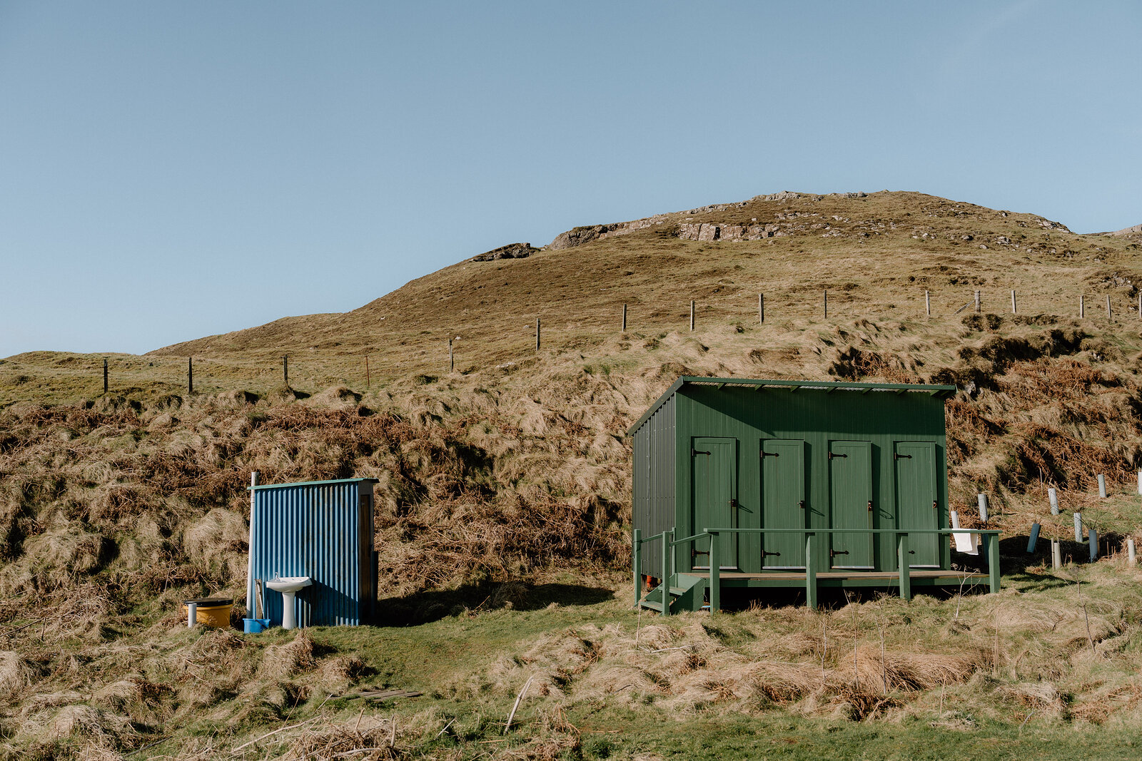 A green shed and a blue water tank sit at the foot of a grassy hill on the Isle of Canna.