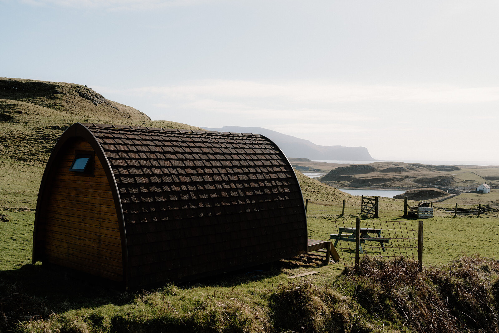 A dark timber camping pod sits on a grassy hillside above a bay on the Isle of Canna.
