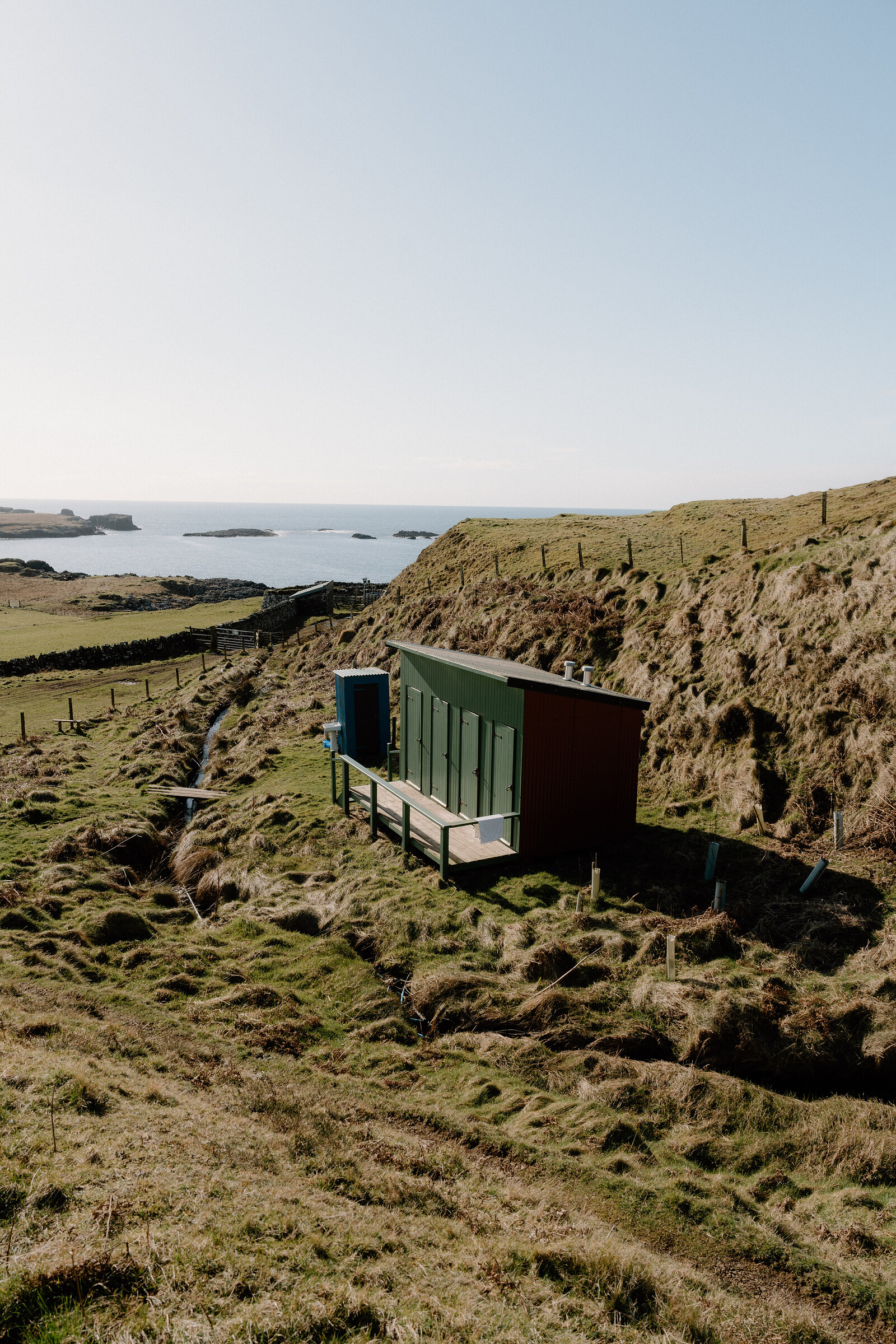 A green and dark timber cabin sits on a grassy slope above a calm bay on the Isle of Canna.