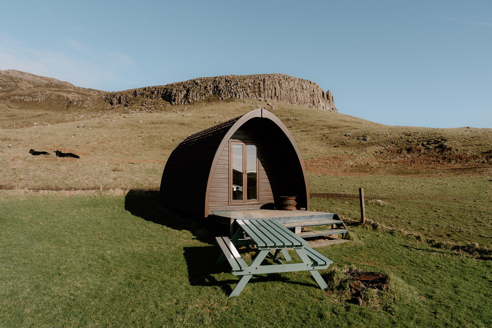 A camping pod and picnic table sit on grass beneath a rocky hill on the Isle of Canna.