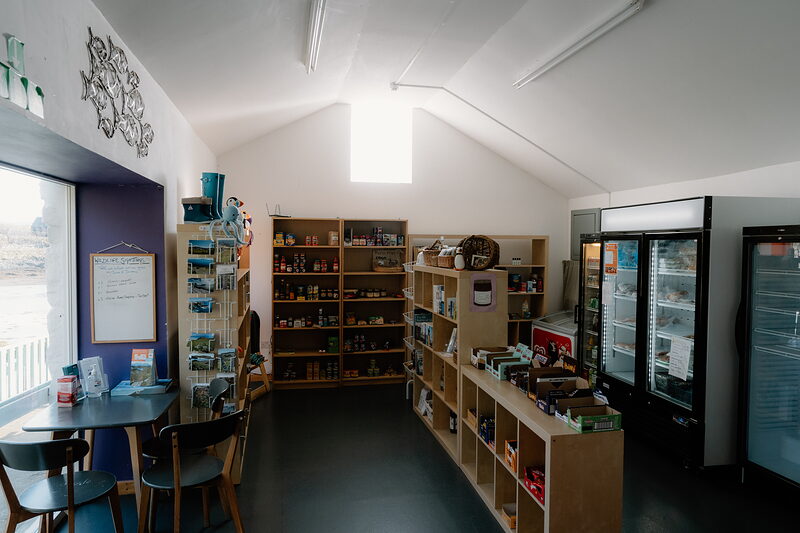 Inside the community shop: shelves of groceries, cards and a cold cabinet, a table by the window.