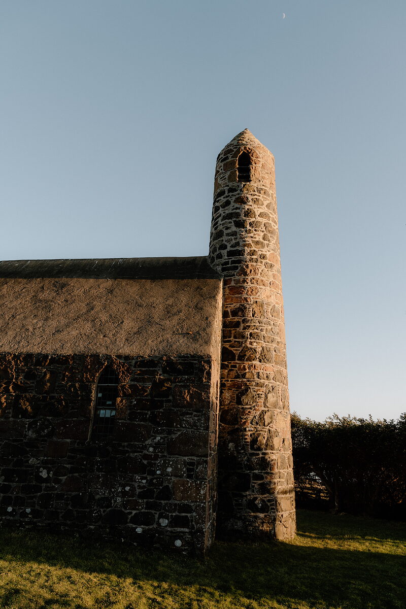 A round bell tower and thatched chapel catch low sun on the Isle of Canna, a pale moon overhead.
