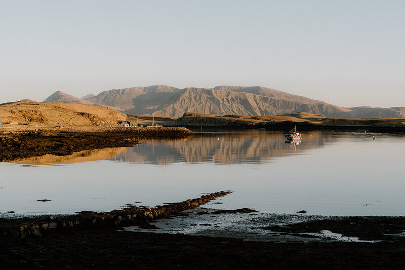 A small boat sits in a mirror-calm bay below layered hills on the Isle of Canna, a cottage on the shore.