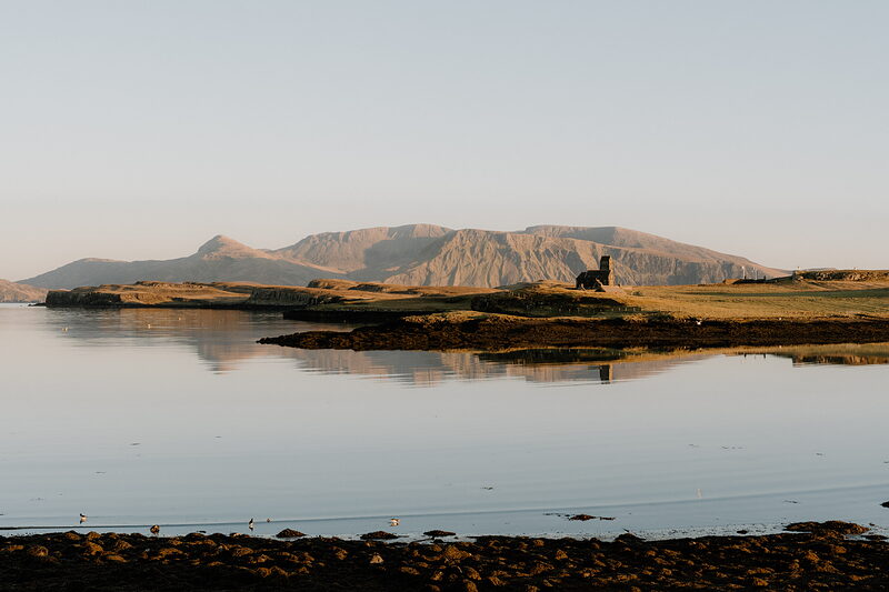 A ruined stone tower stands on a grassy headland, mirrored in a calm bay on the Isle of Canna.