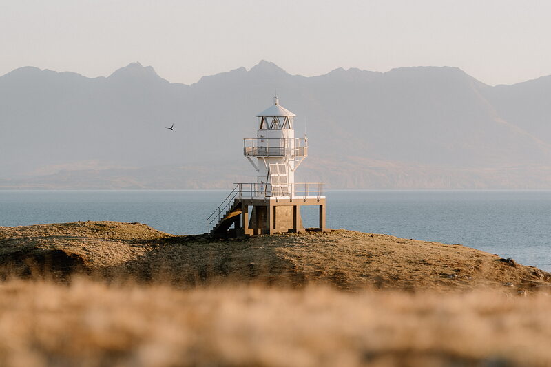 A white lighthouse tower with a stairway stands on a grassy headland on the Isle of Canna, mountains rising behind.