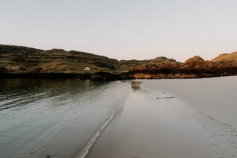 A sandy beach curves beneath grassy hills on the Isle of Canna at golden hour, a white cottage visible on the shore.