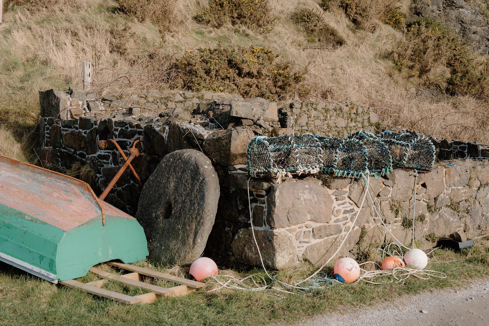 A millstone, coiled ropes and buoys sit beside a stone wall on the Isle of Canna, a green boat parked nearby.