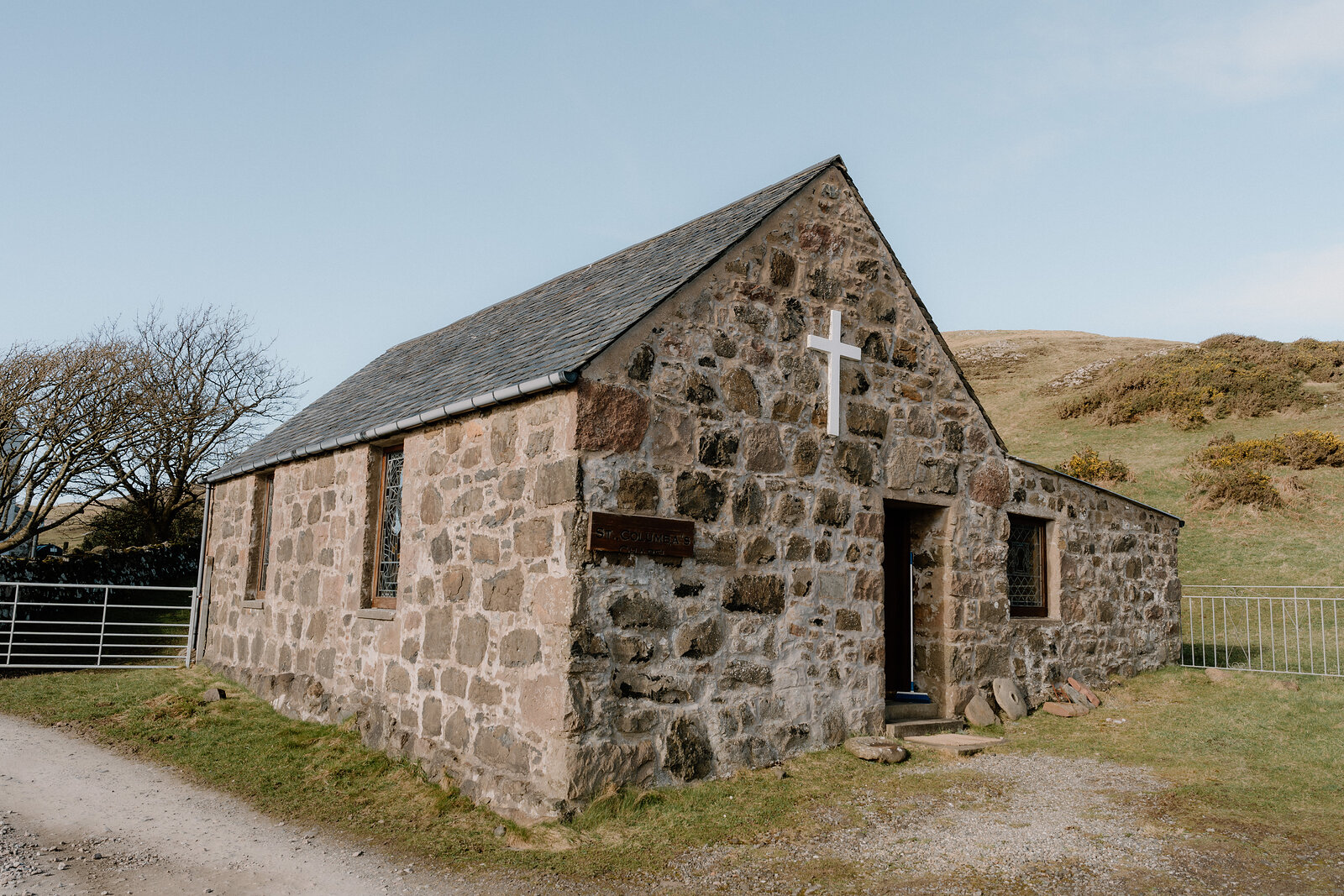 A small stone chapel with a white cross stands beside a metal gate on the Isle of Canna.