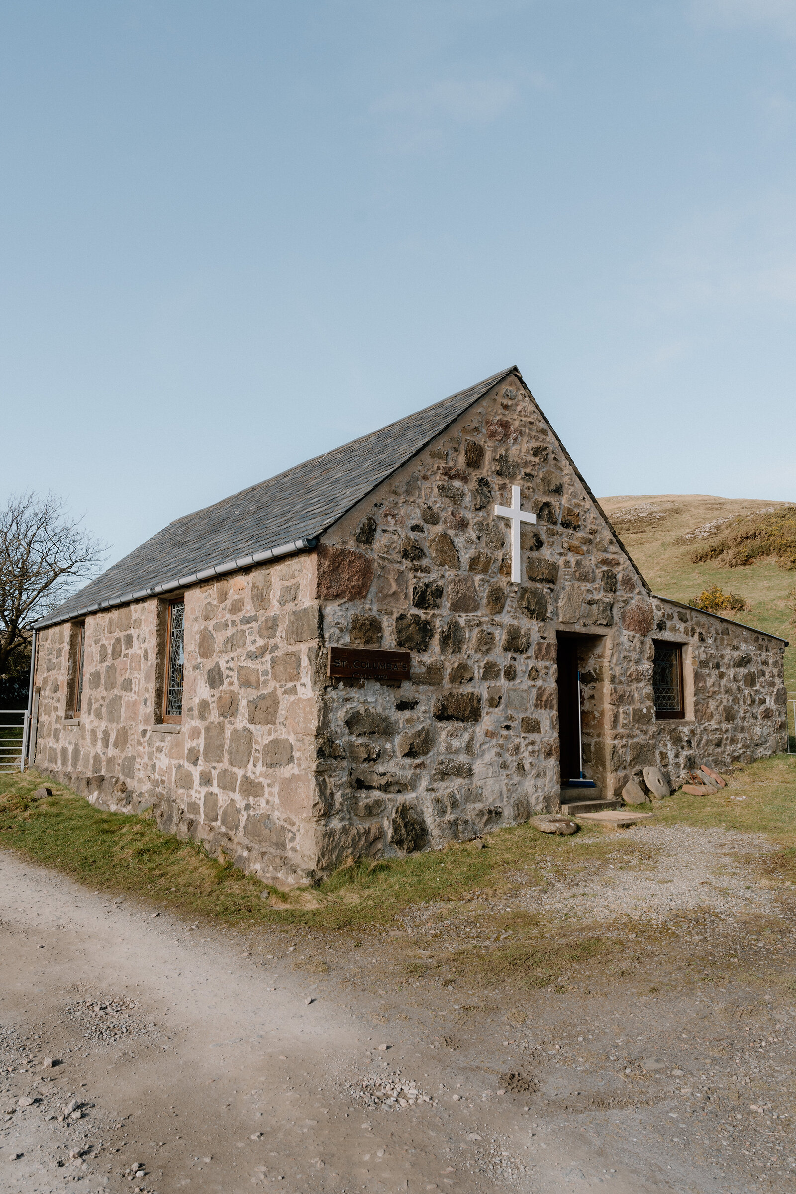 A small stone chapel with a white cross and pitched slate roof stands on a gravel path on the Isle of Canna.
