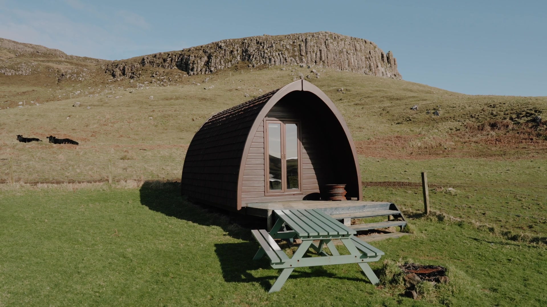 A dark timber glamping pod with an arched roof and picnic bench sits on a grassy slope below a basalt-columned cliff, cattle grazing in the field beyond.