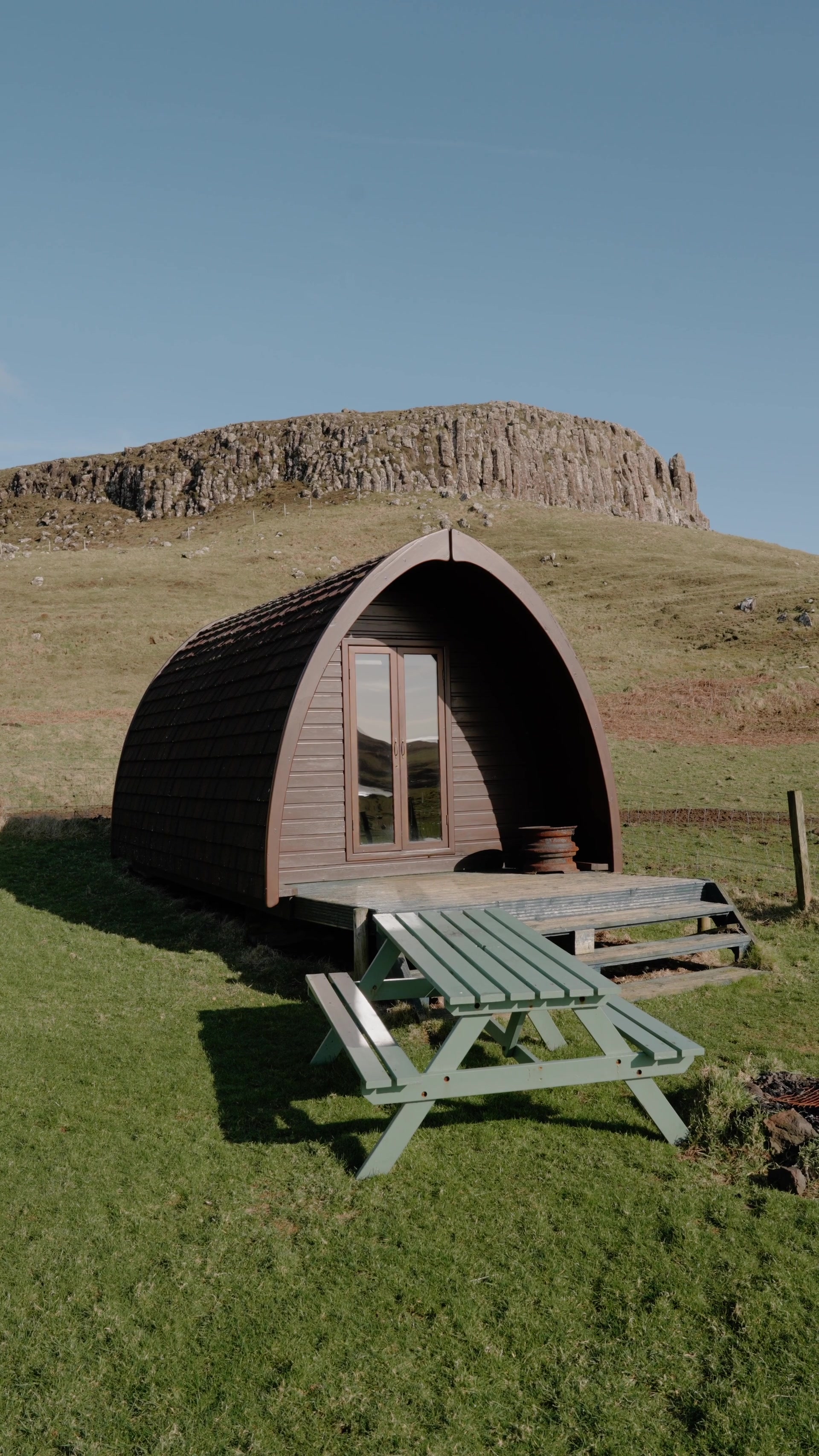 A dark timber glamping pod with an arched shingled roof and glass double doors sits on a grassy slope, a green picnic bench in front and a rocky, columned escarpment rising behind.