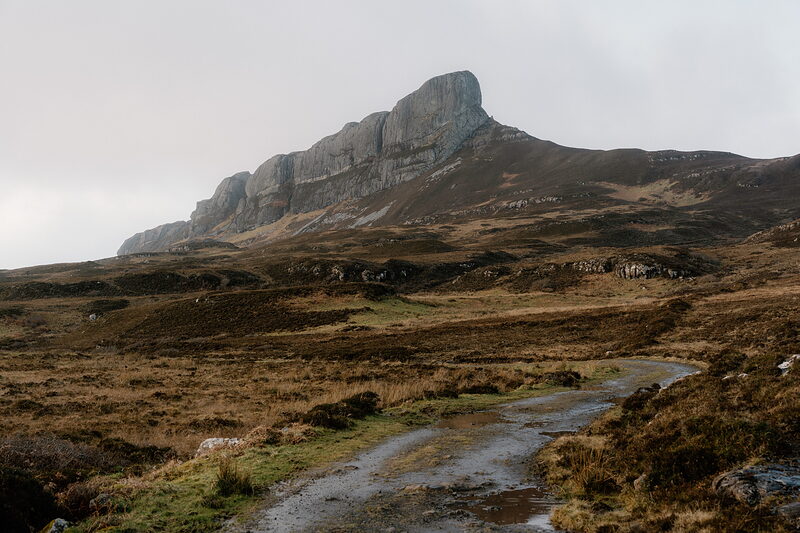 A jagged rocky peak rising above moorland, seen from a path across the hillside.