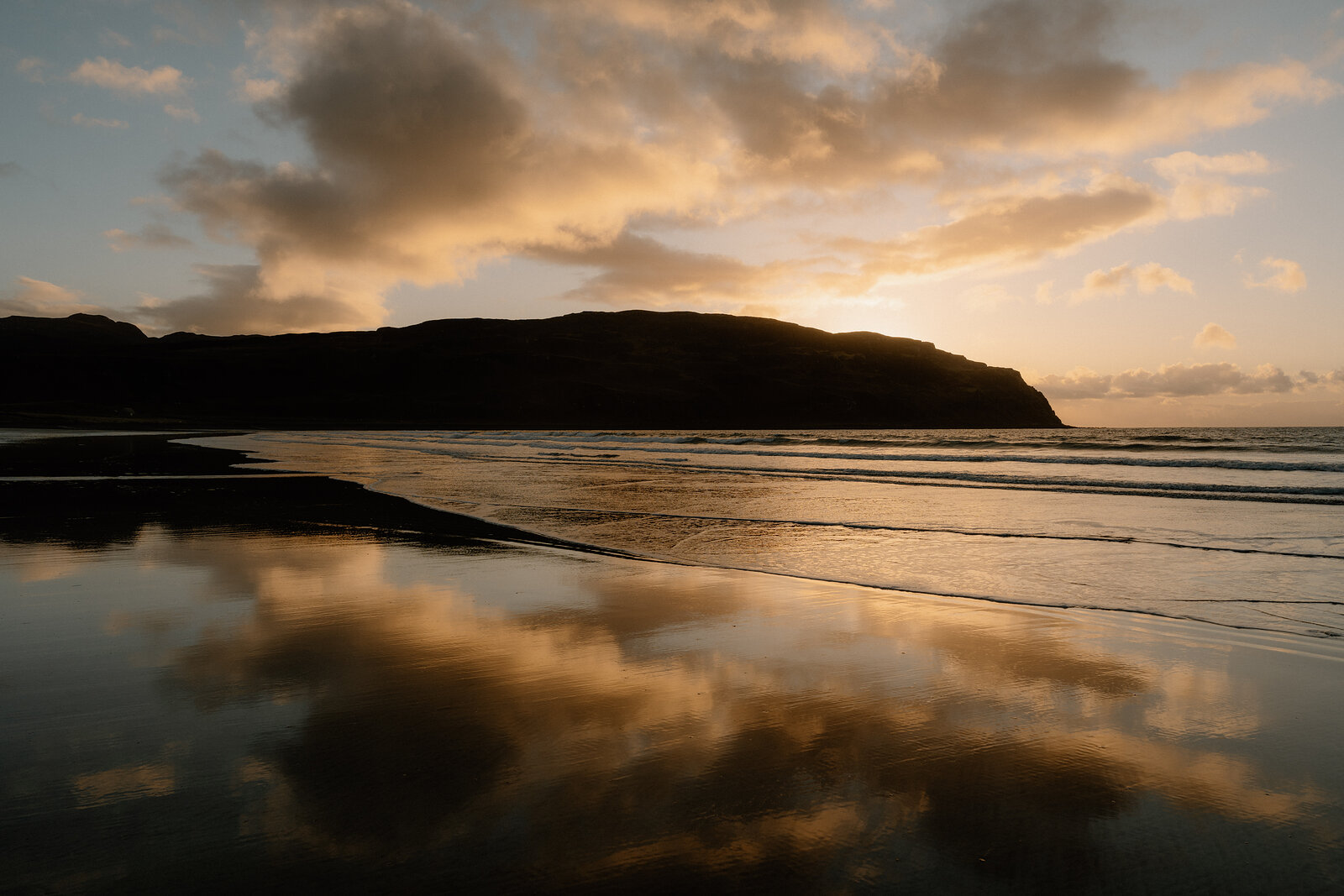 Waves reflecting a golden sunset beneath a silhouetted headland on the beach
