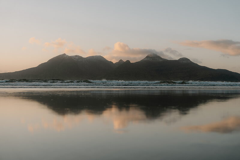A silhouetted mountain range reflected in the still water of the beach at dusk.