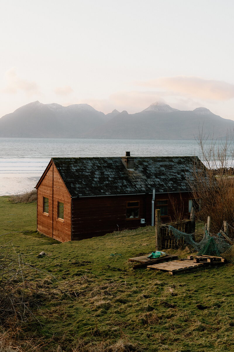 A timber cottage beside the shore, mountains rising across the water behind.