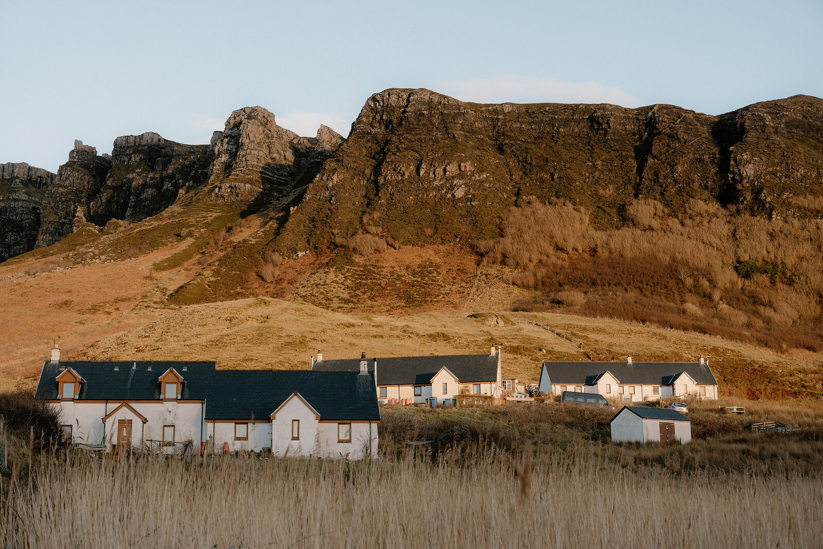 A row of white cottages beneath layered rocky cliffs, evening light catching the grass