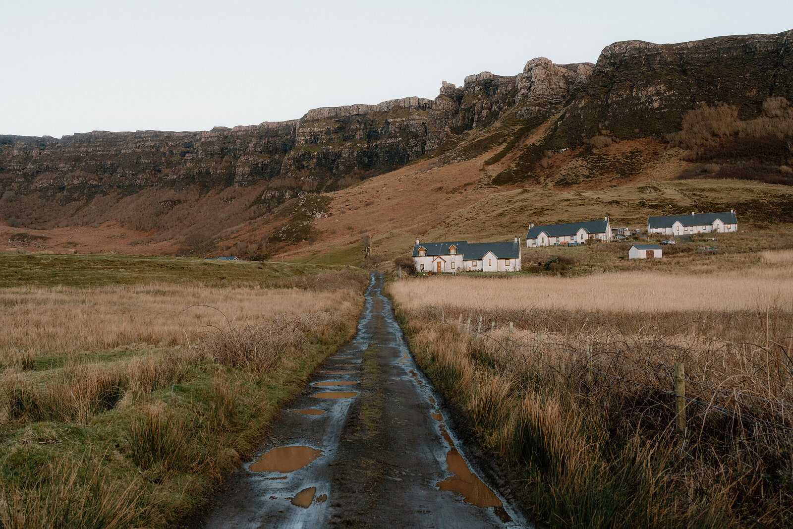 A tarmac lane leading towards a row of white cottages beneath layered rocky cliffs