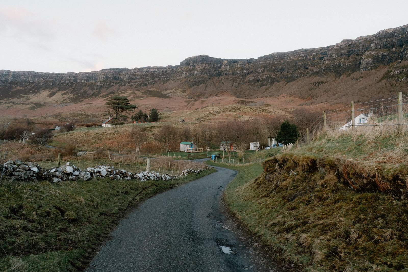 A tarmac lane winding past a stone wall and cottages beneath layered rocky cliffs