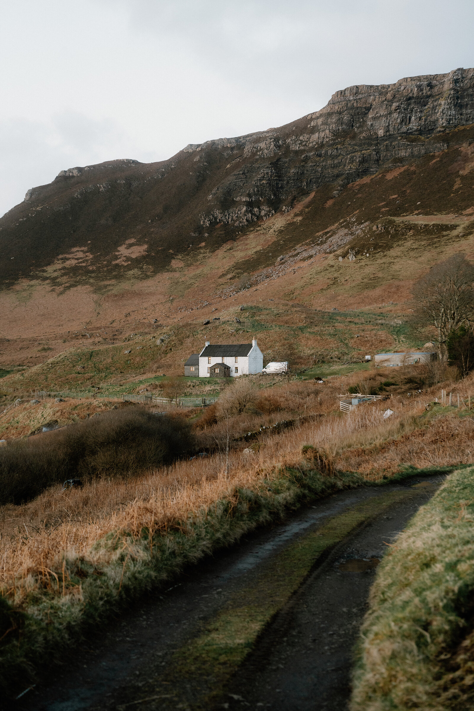 A white cottage set below layered rocky cliffs, a puddled track leading towards it
