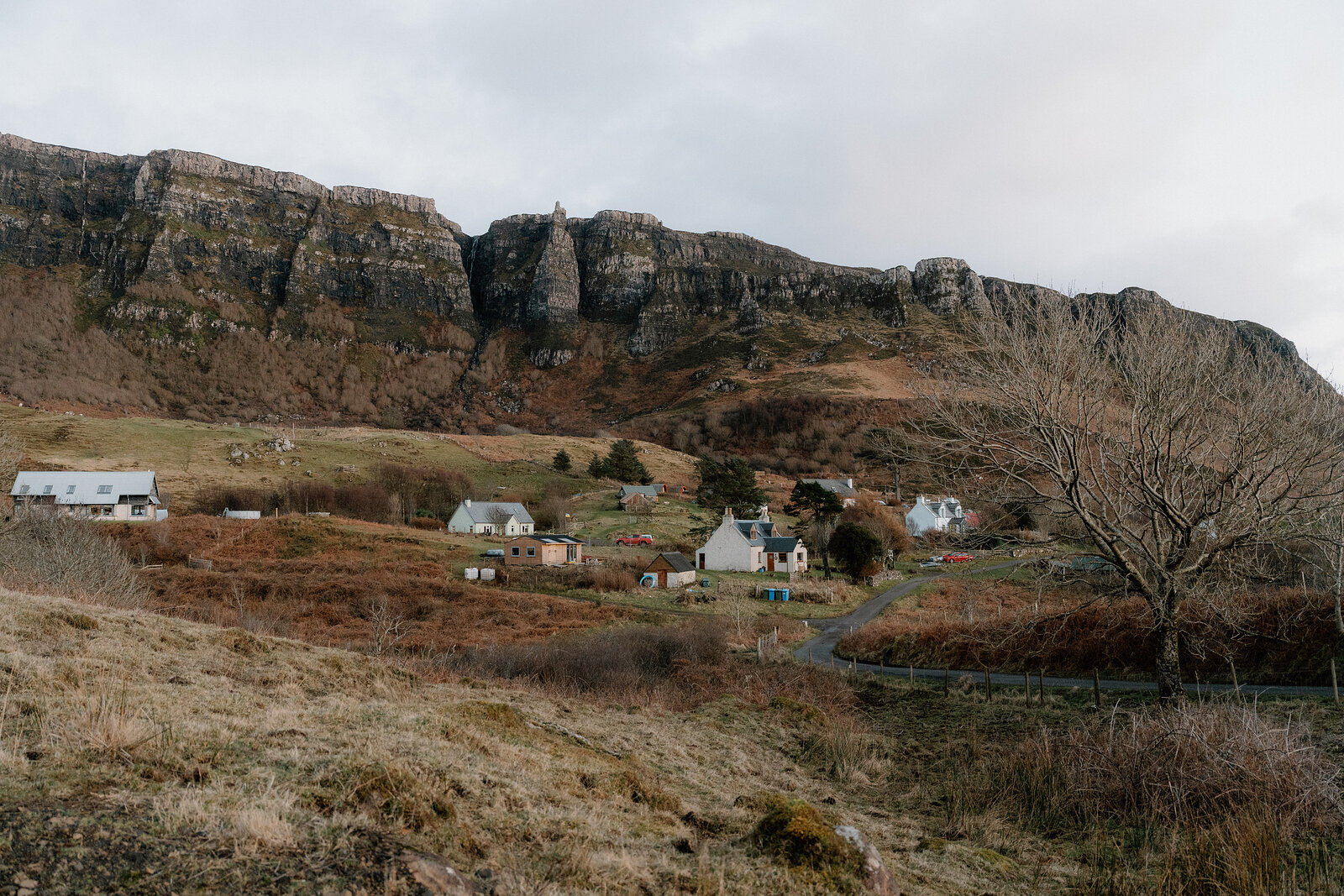 A scatter of white cottages beneath layered rocky cliffs, a bare tree standing in the foreground