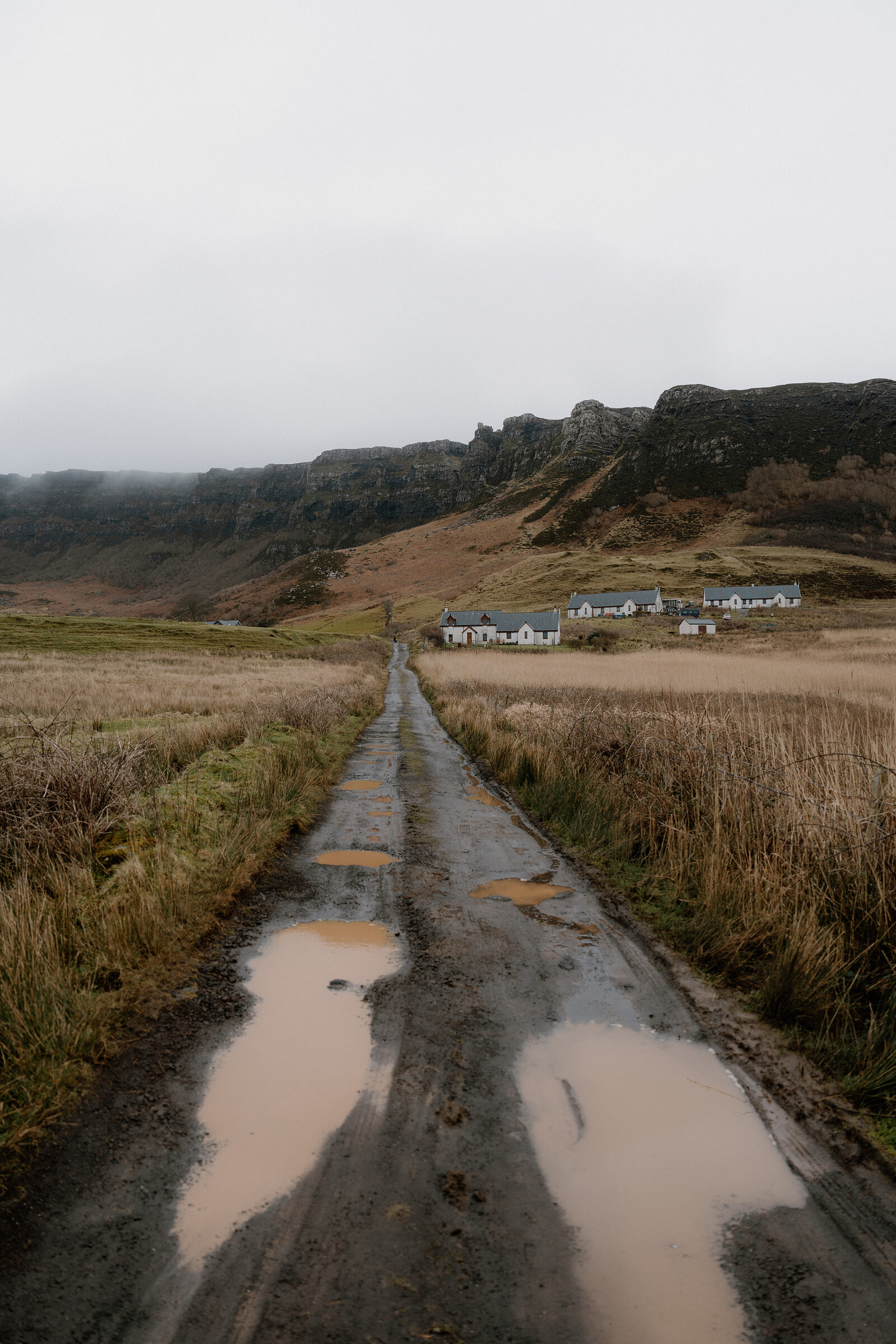 A puddled track leading towards white cottages beneath layered rocky cliffs