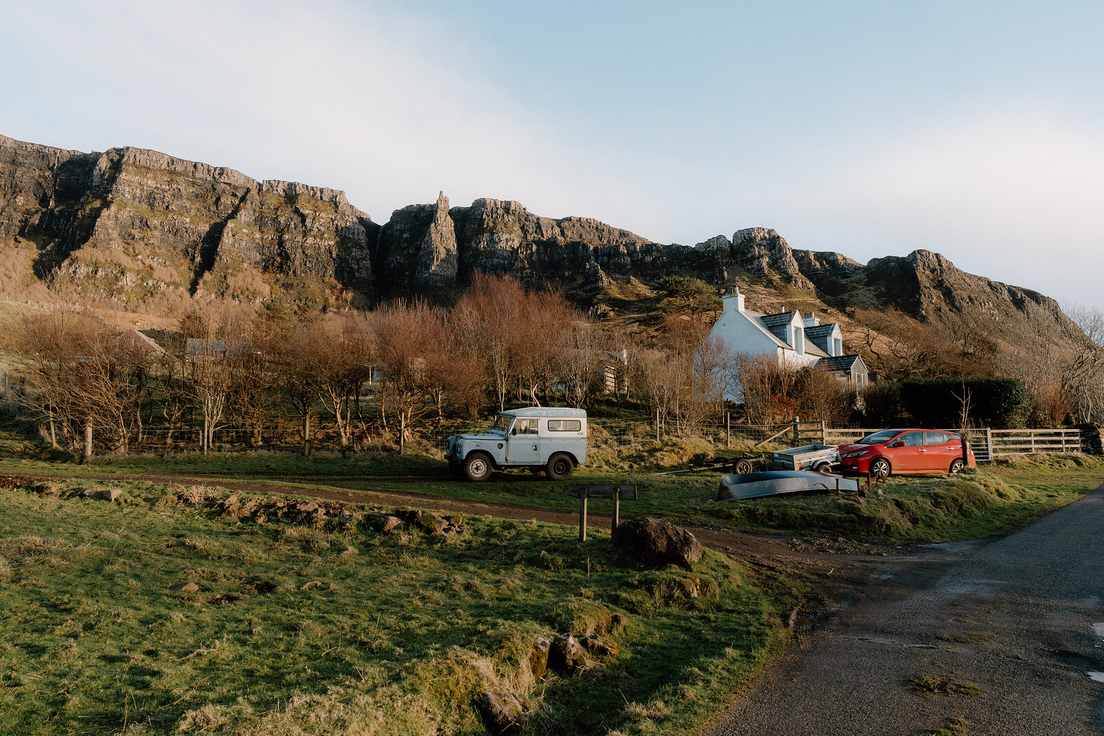 A vintage Land Rover parked beside a white cottage below layered rocky cliffs