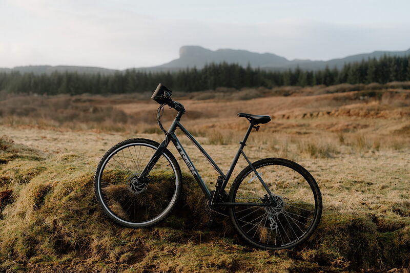 A hire bike leaning on a grassy bank on the moor, the ridge of An Sgùrr beyond a conifer wood.