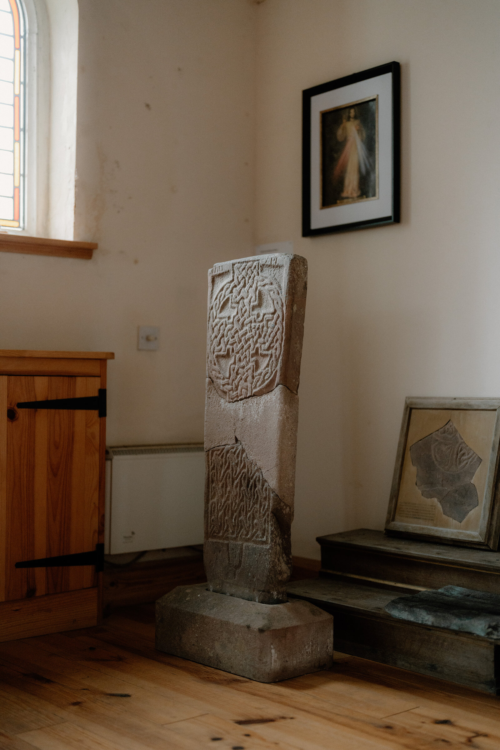 A carved stone cross-slab stands in the corner of a small church beside a wooden cabinet.