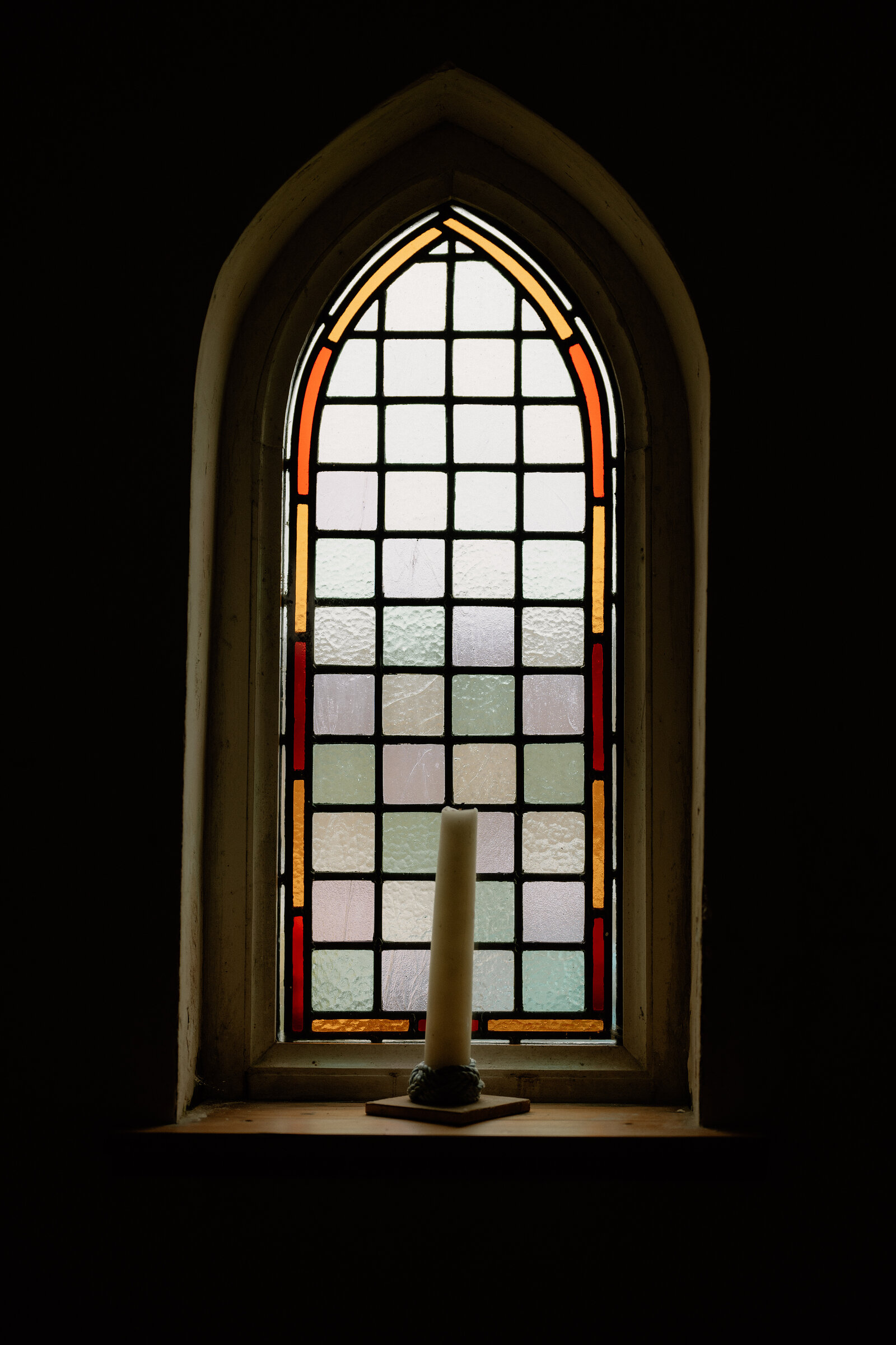 A candle sits on the sill of a stained-glass arched church window.