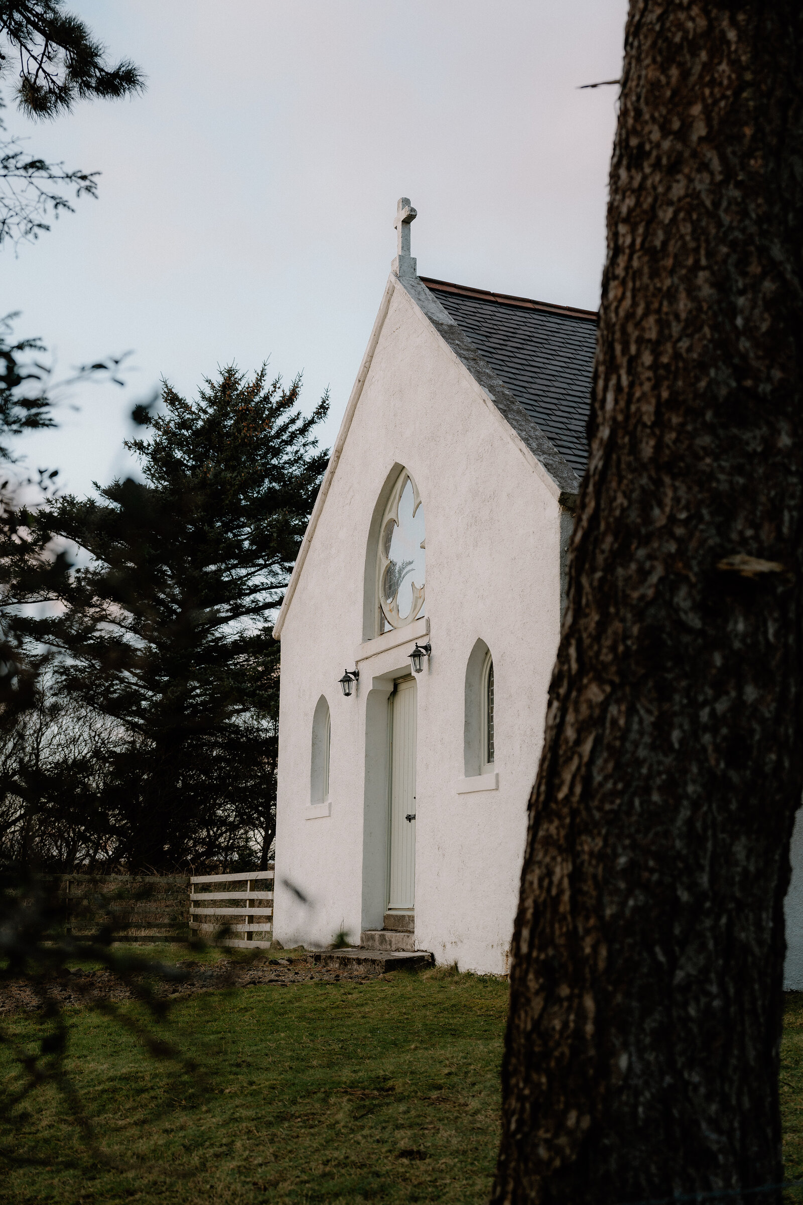 A small whitewashed church with a quatrefoil window stands beneath a cross-topped gable.