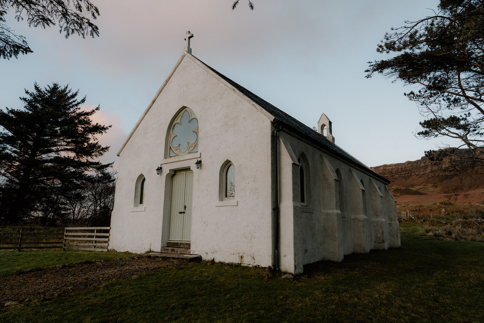 A small whitewashed church with a bell cote stands before rocky hills.