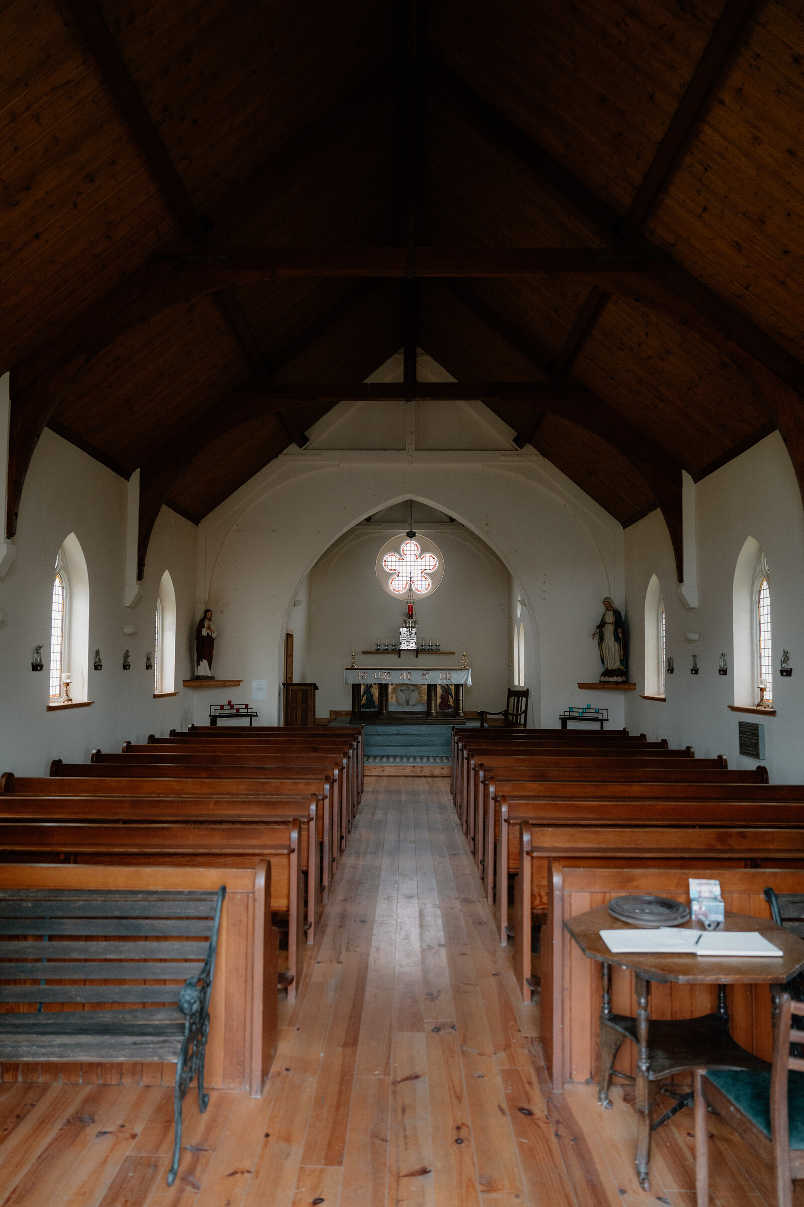 Wooden pews line a church nave beneath a beamed ceiling and quatrefoil window.