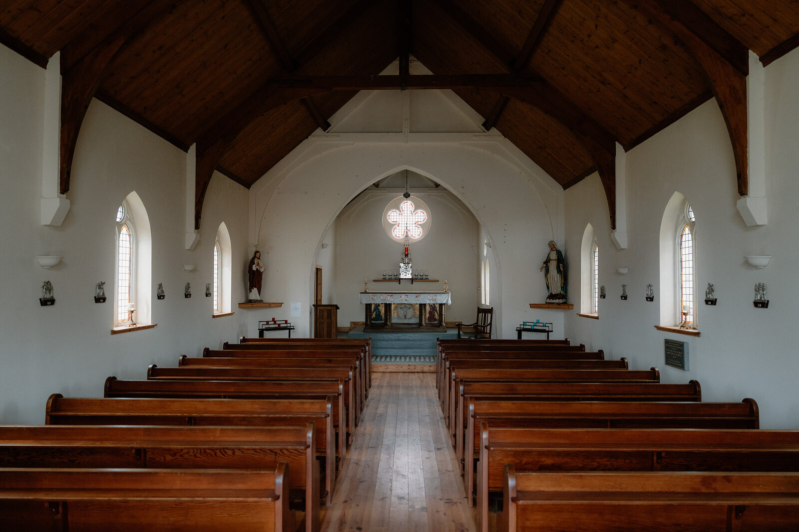 Wooden pews line a church nave leading to an altar lit by a quatrefoil window.