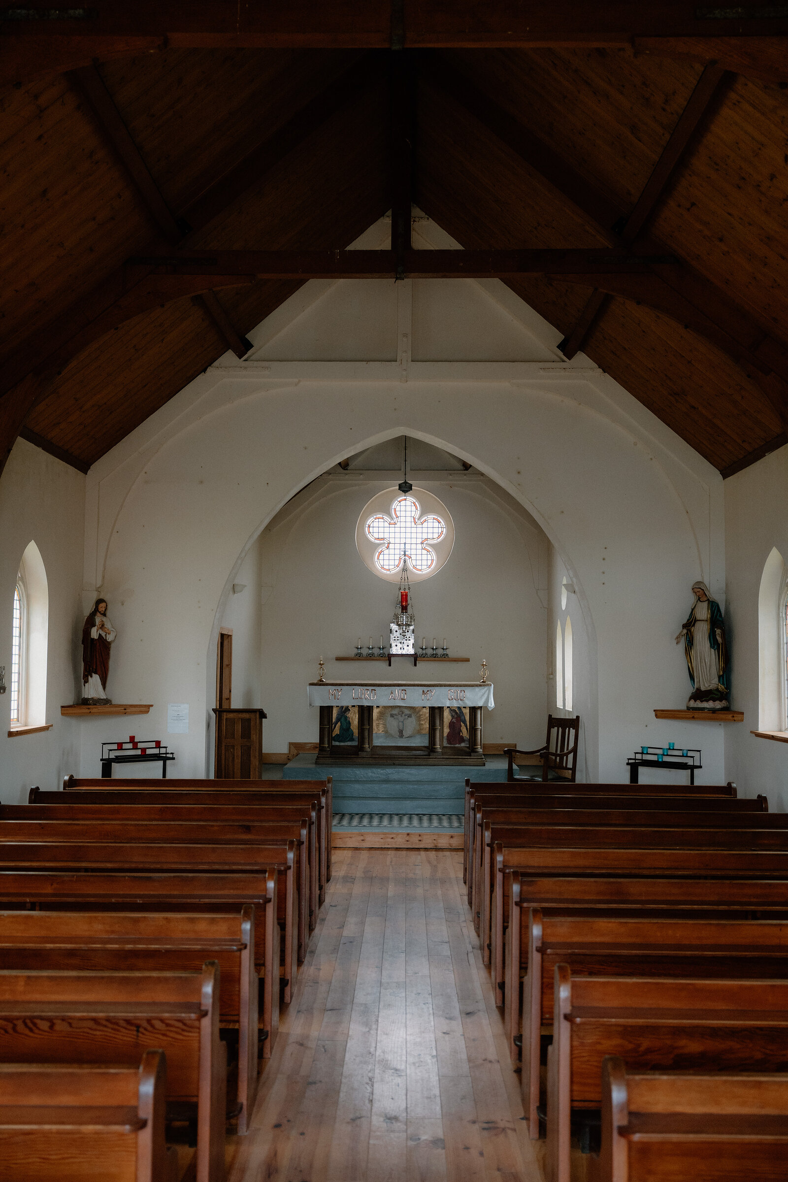 Small statues flank a stained-glass arched window above a church's wooden pews.