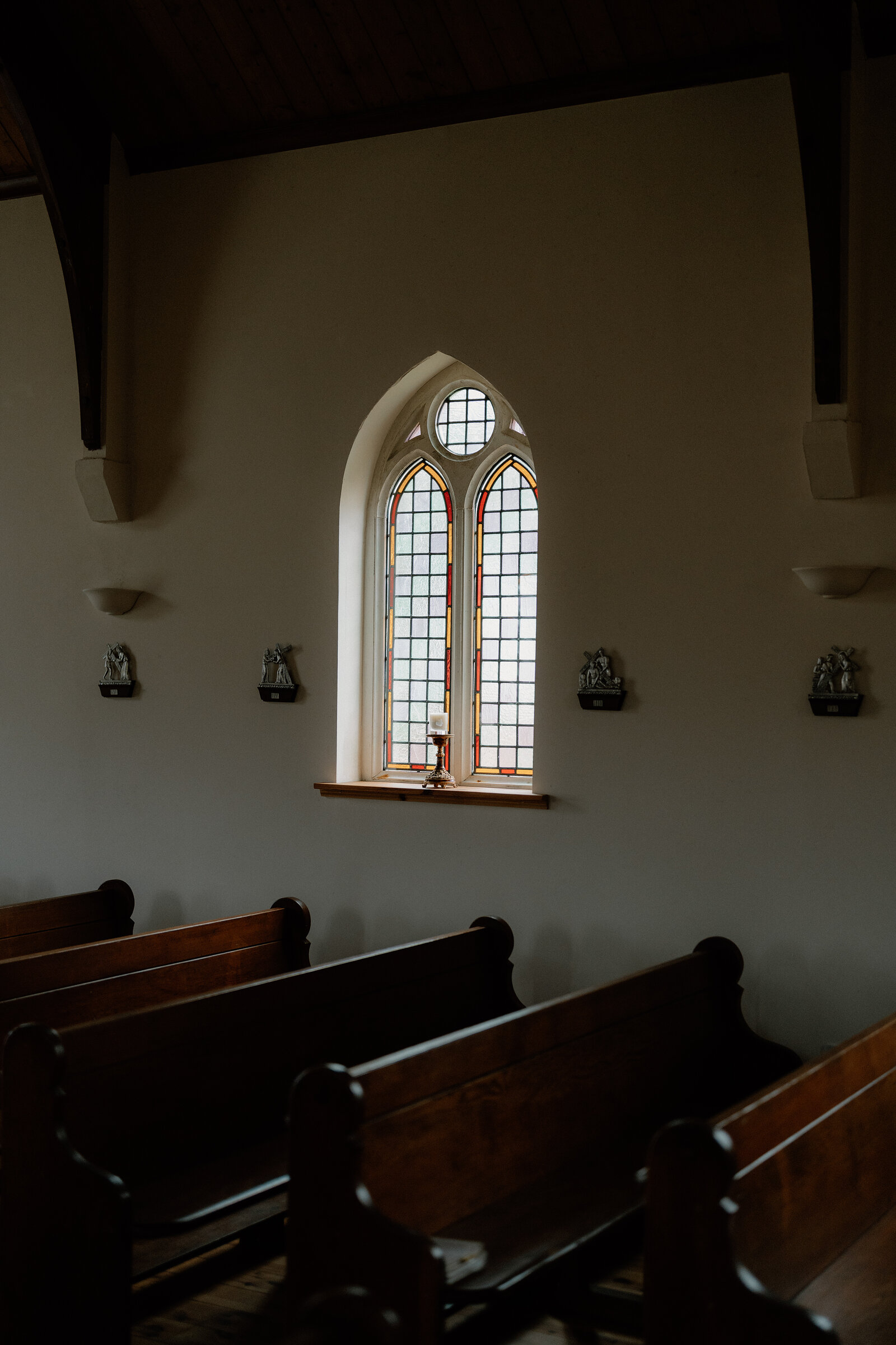 A stained-glass arched window glows above a church's dark wooden pews.