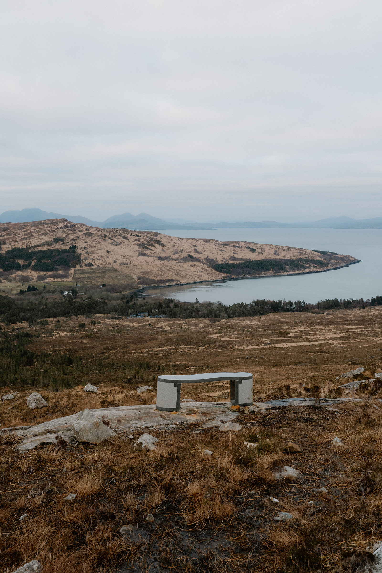 A stone bench sits on a hilltop above a wooded bay on the Isle of Rum.